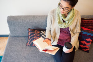 Person wearing a scarf sitting on a grey couch reading a book and holding a cup of coffee