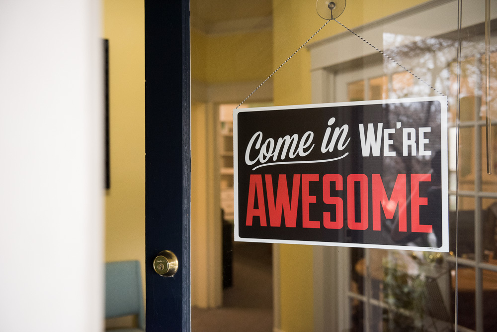 Welcome sign hanging from post that says “Come In, We’re Awesome”