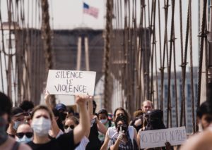 Multiracial group of protesters walks over a bridge. In image center, one protester holds up a white sign with SILENCE IS VIOLENCE in black letters.
