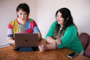 Two women sit in front of a laptop at a wooden table, discussing details of a creative consultation.