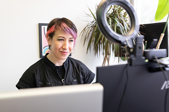 Sarah sits at her desk with two computers in front of her and a ring light pointed in her direction.